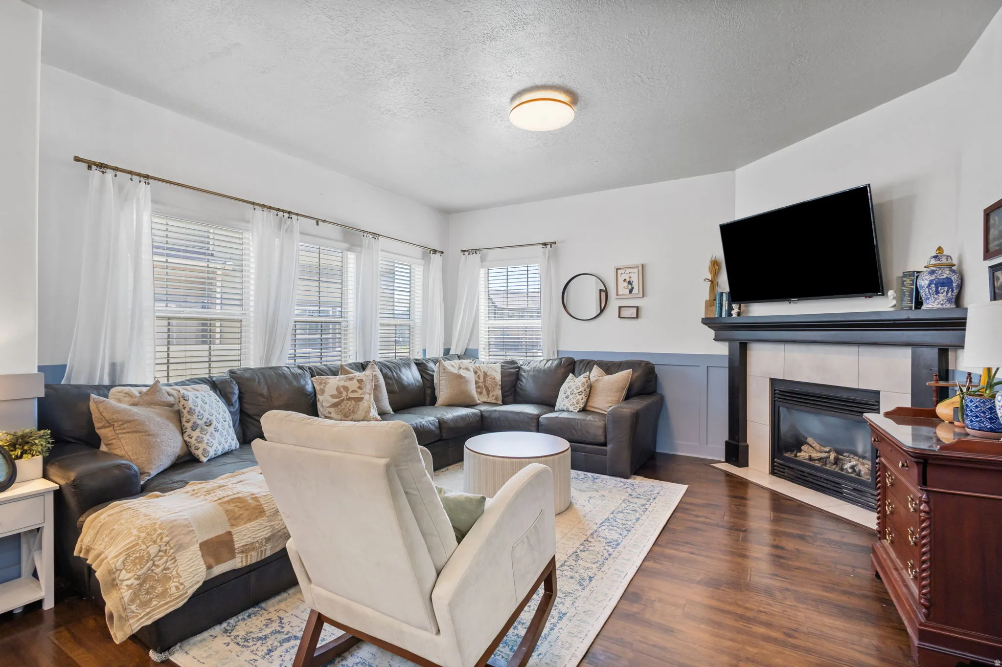 Living area featuring rich wood-finish flooring, a corner-set fireplace with a dark mantel and light tile surround, and multiple windows with blinds and sheer drapes