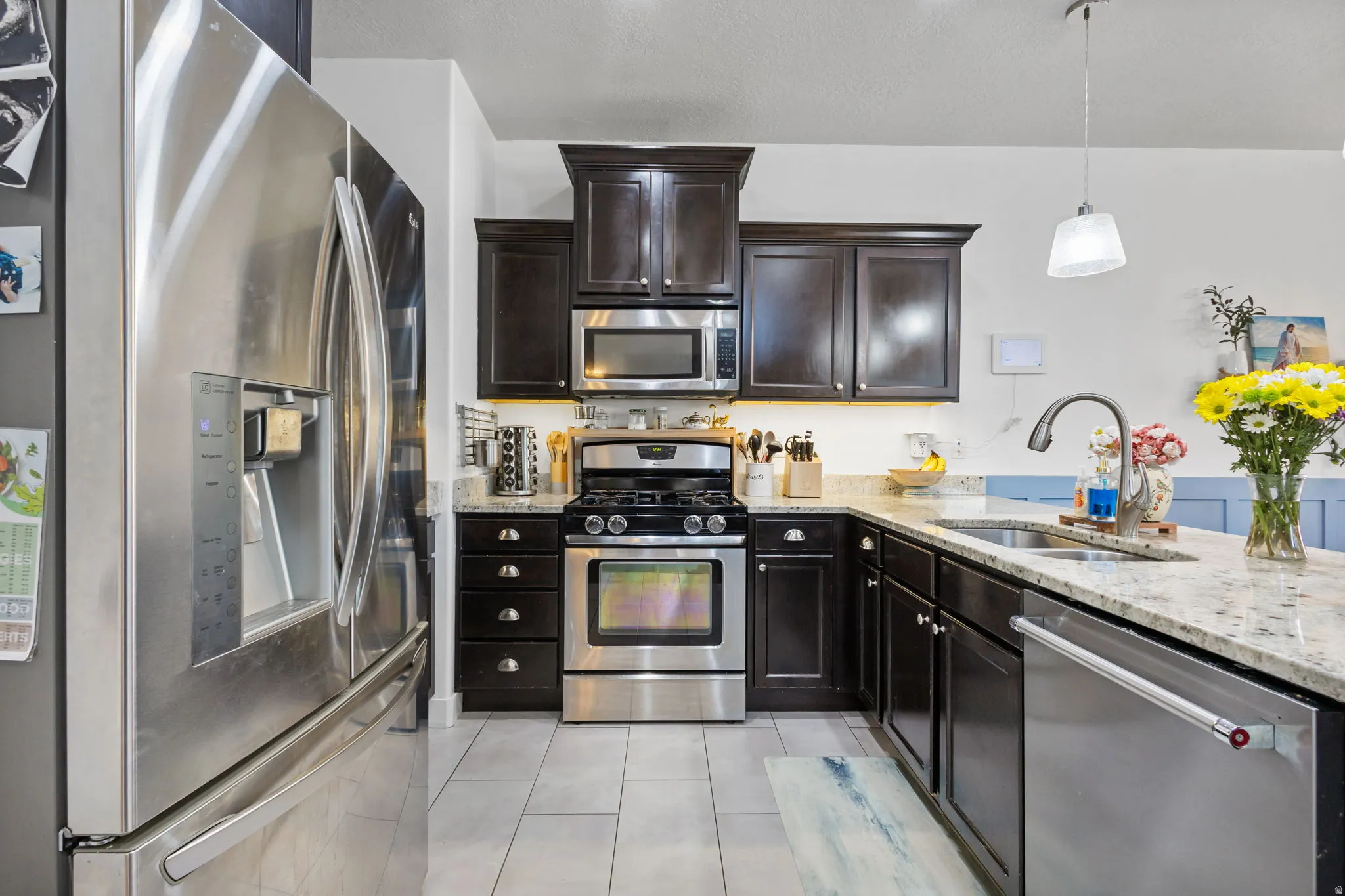 Contemporary kitchen featuring dark wood cabinetry, stainless steel appliances, light granite countertops, and light-toned tile flooring