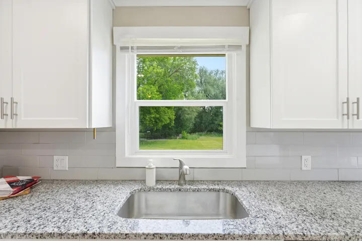 Kitchen sink area featuring an undermount stainless steel sink, speckled granite countertop, white shaker cabinetry with brushed nickel hardware, and a light gray subway tile backsplash
