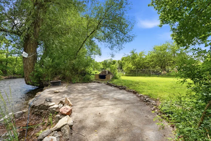 Riverside concrete patio with a built-in outdoor grill, bordered by mature trees and a manicured lawn