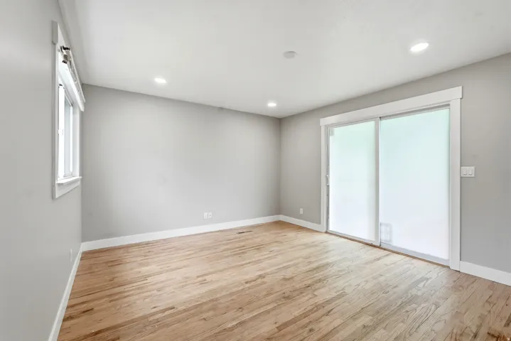Spacious interior room featuring wood-finish flooring, light gray walls, white trim, recessed lighting, a large window, and a sliding glass door