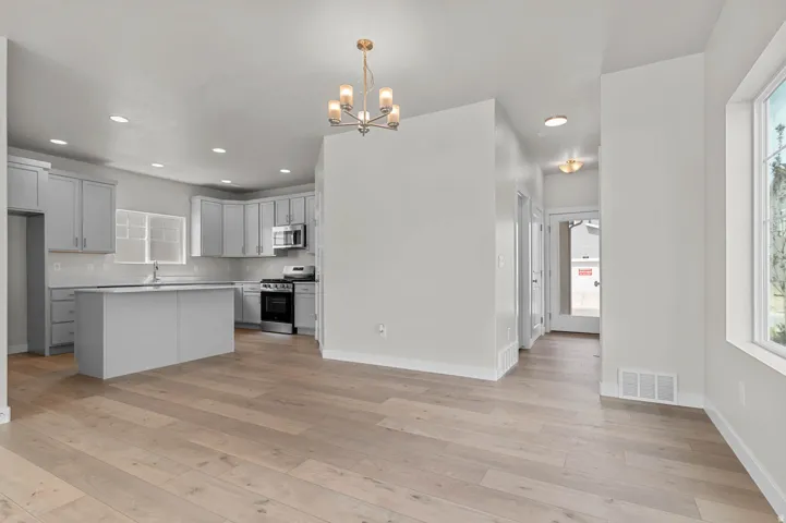 Open-concept living space with light wood-finish flooring, featuring a kitchen with gray cabinetry, a central island, and stainless steel appliances, complemented by recessed lighting and a decorative chandelier