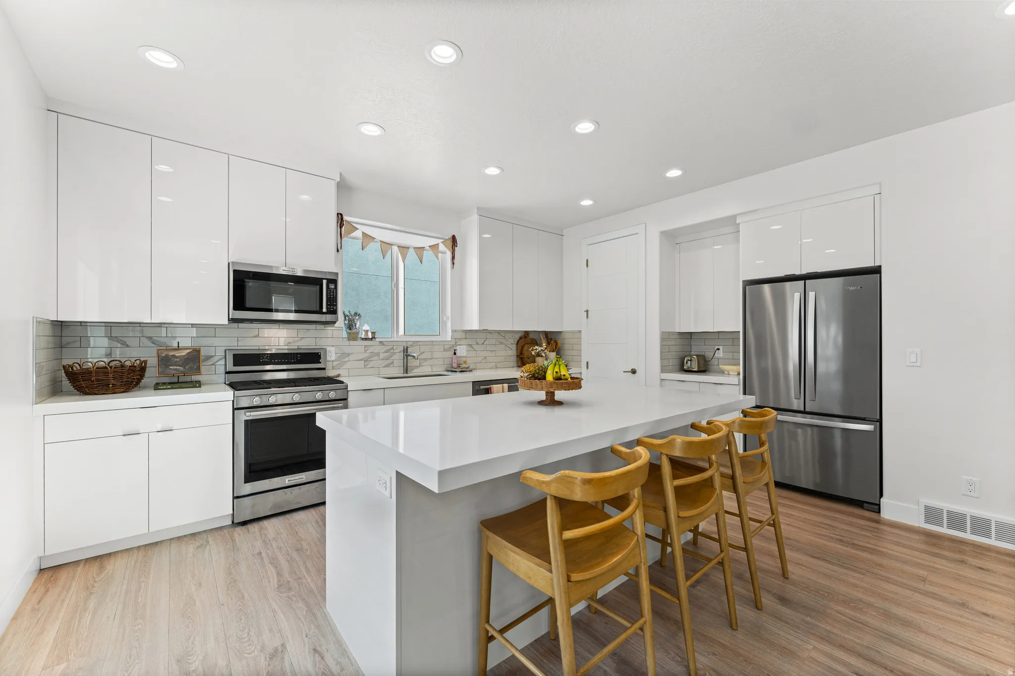 Contemporary kitchen featuring high-gloss white cabinetry, stainless steel appliances, a white waterfall island, wood-finish flooring, and recessed lighting
