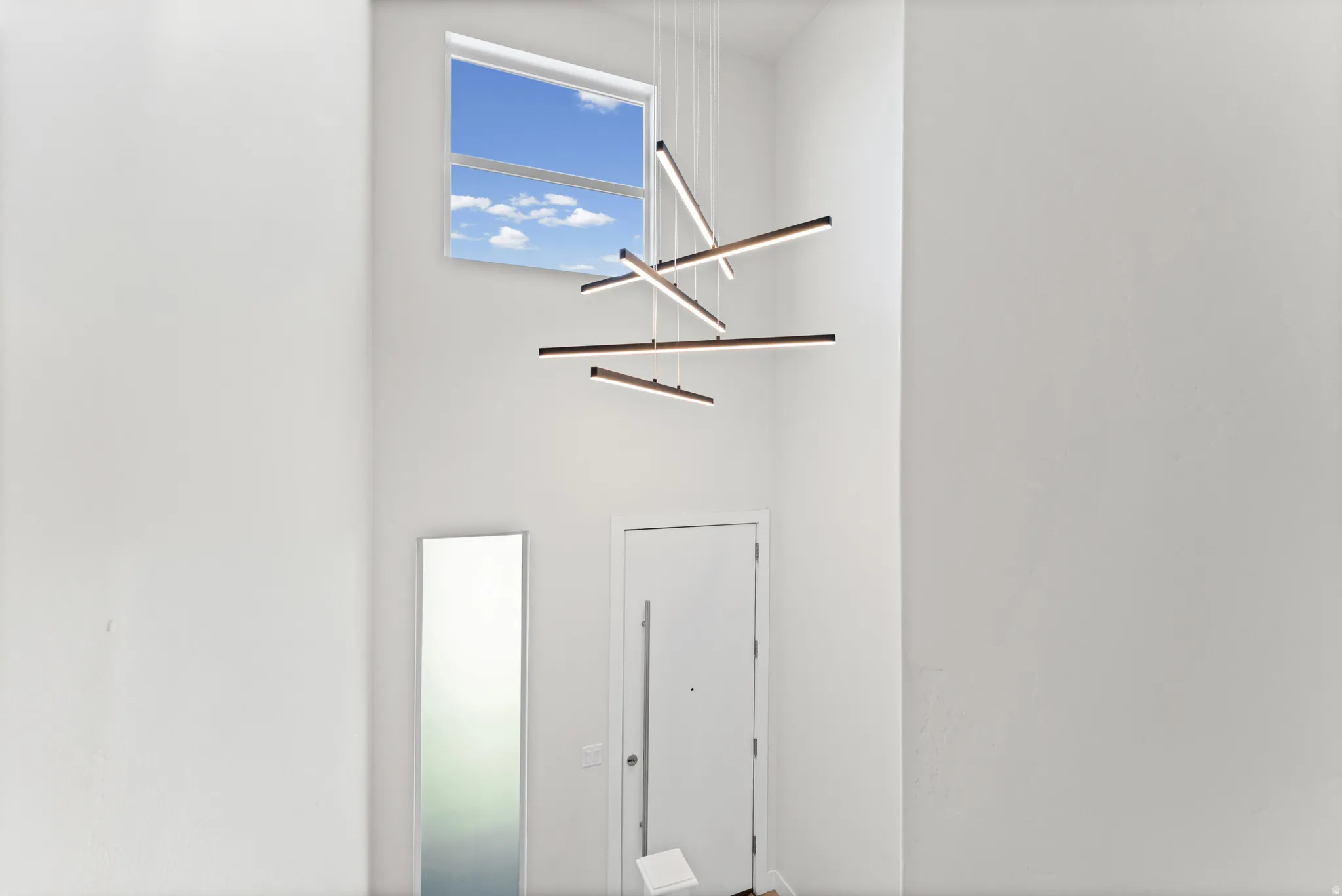 Two-story entry featuring high ceilings, a contemporary linear chandelier, a transom window, and a white entry door with a horizontal pull handle