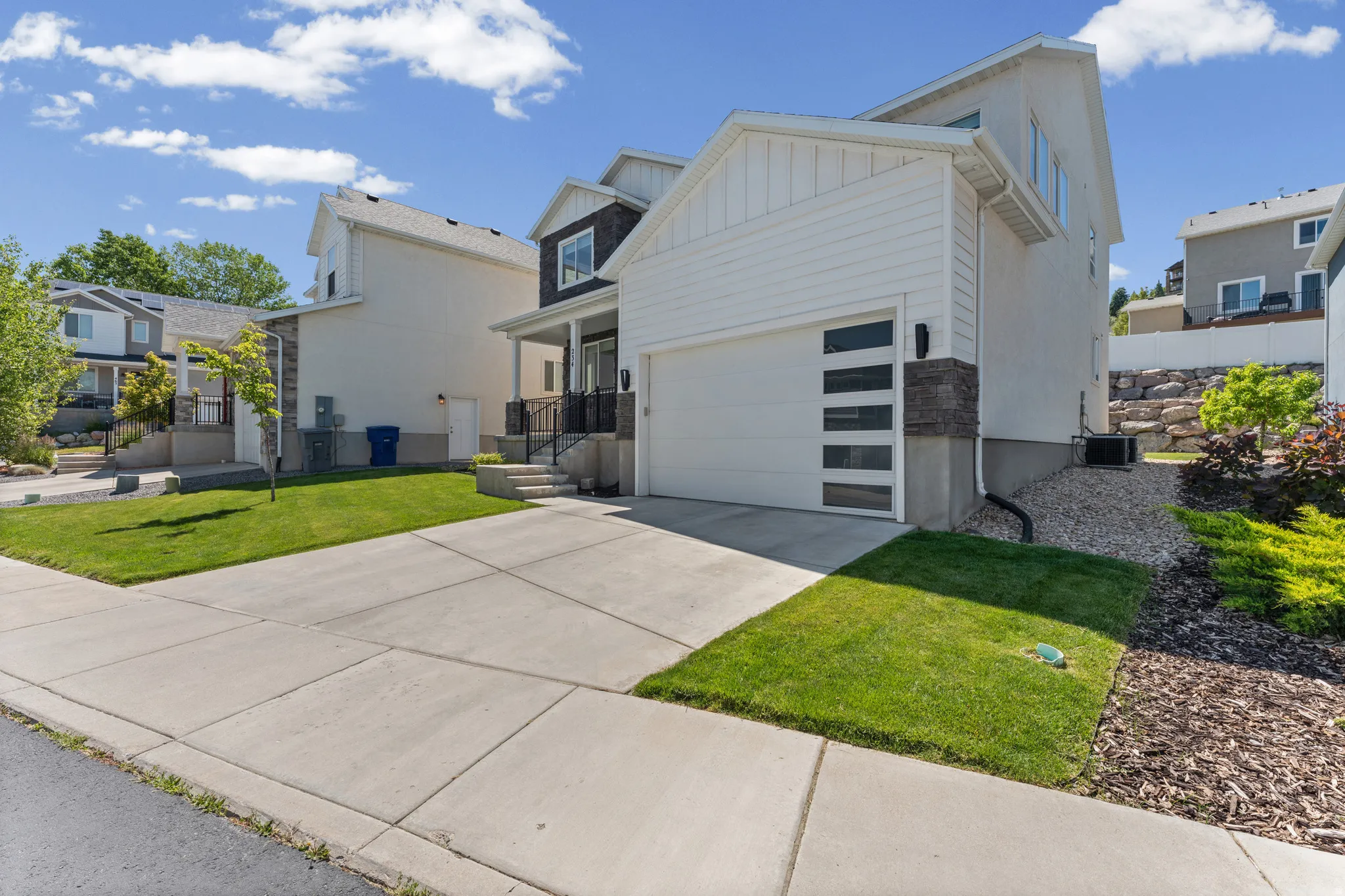 Modern architectural style featuring a clean white facade with dark accent siding, a contemporary garage door with horizontal windows, a concrete driveway, and front landscaping with turf and mature plantings