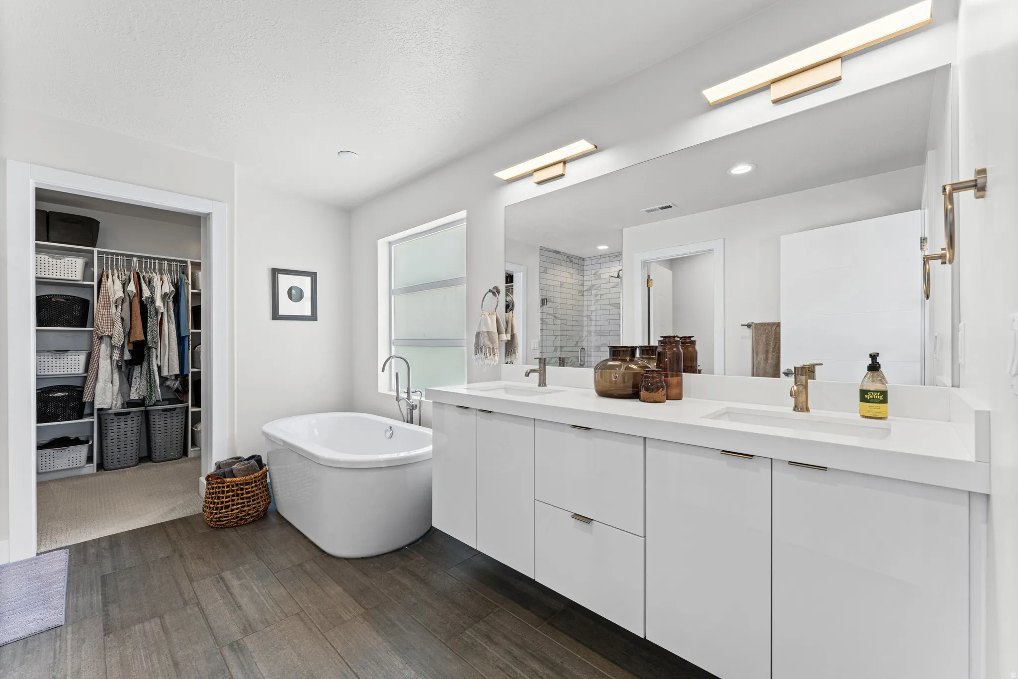 Contemporary bathroom featuring a freestanding soaking tub, wood-finish flooring, a double vanity with white cabinetry, and a large wall-mounted mirror with integrated lighting