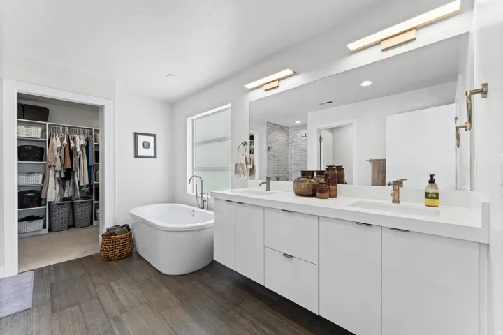 Contemporary bathroom featuring a freestanding soaking tub, wood-finish flooring, a double vanity with white cabinetry, and a large wall-mounted mirror with integrated lighting