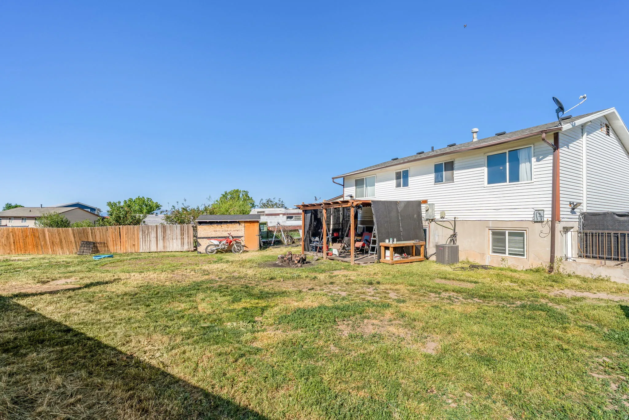 Expansive backyard featuring a large grassy area, a wooden privacy fence, a detached shed, a covered patio area, and a white siding exterior on the main residence
