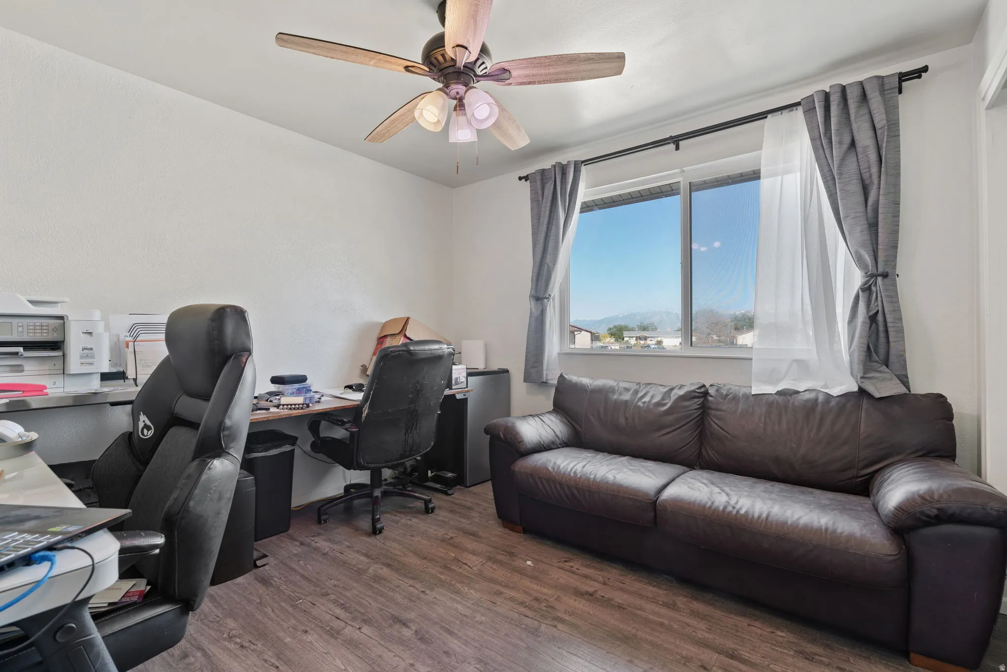 Room featuring wood-finish flooring, a window with exterior scenery, a ceiling fan with light fixture, and white wall finishes