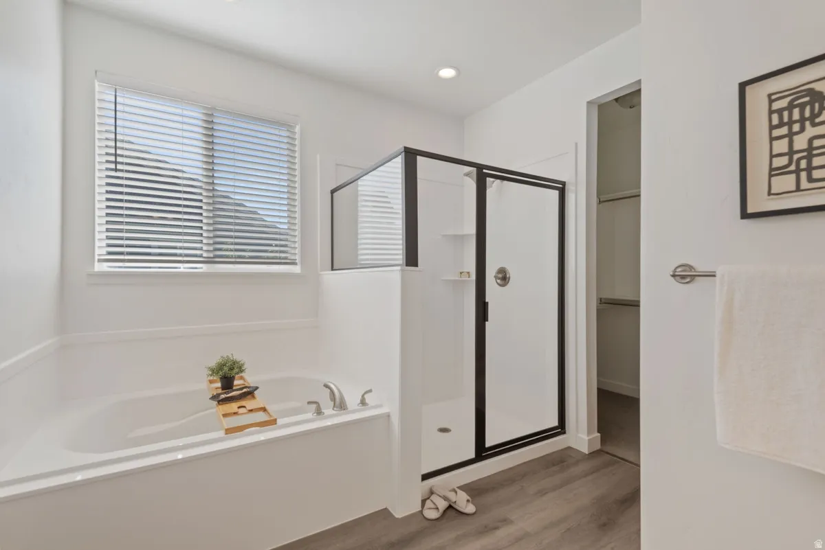 White soaking tub with brushed nickel fixtures, a window with blinds, a separate shower with a black-framed glass enclosure, and wood-finish flooring