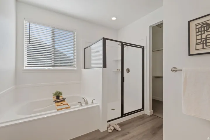 White soaking tub with brushed nickel fixtures, a window with blinds, a separate shower with a black-framed glass enclosure, and wood-finish flooring
