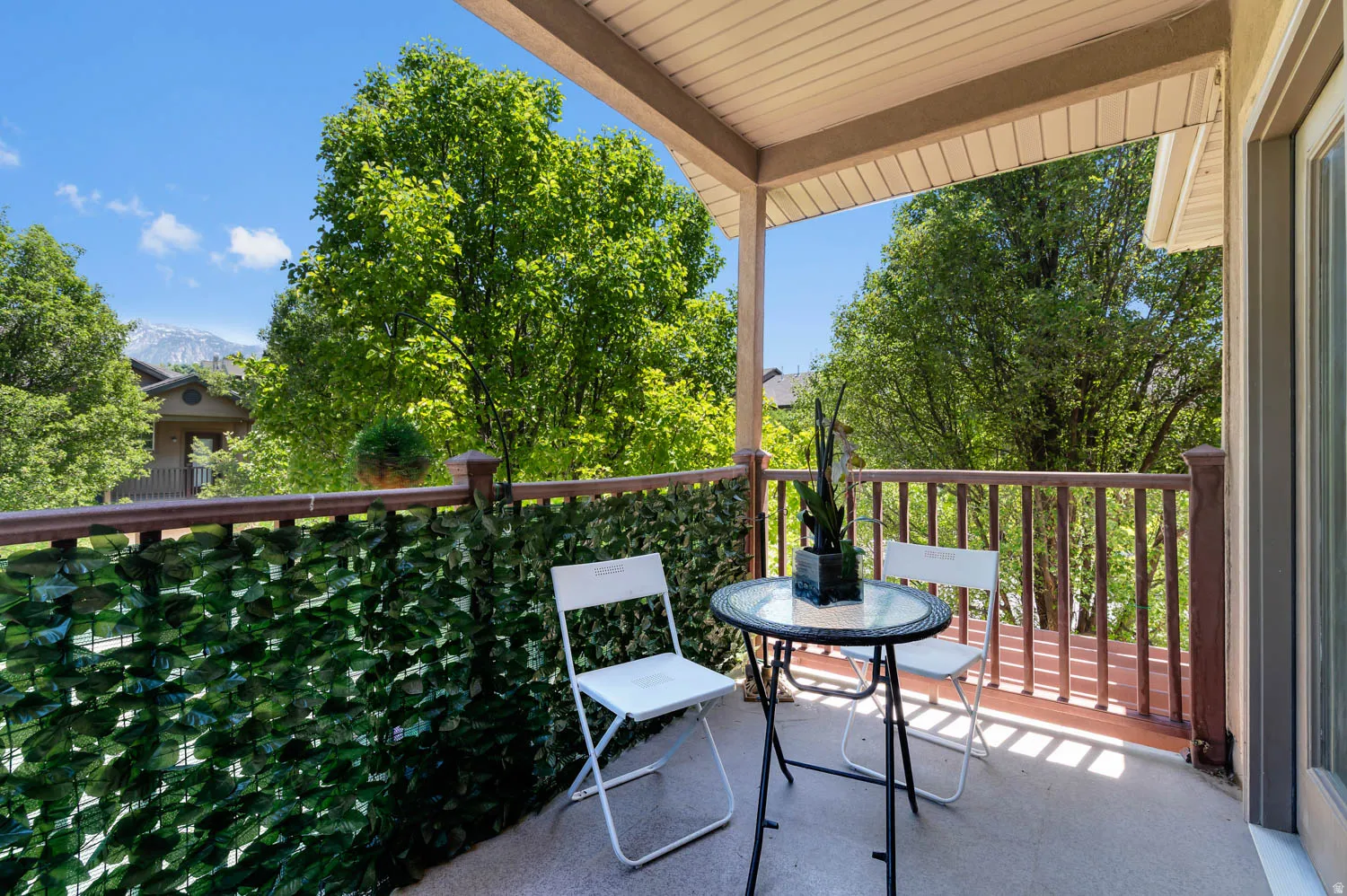 Covered balcony featuring a wood-finish railing, composite decking, and an artificial foliage screen