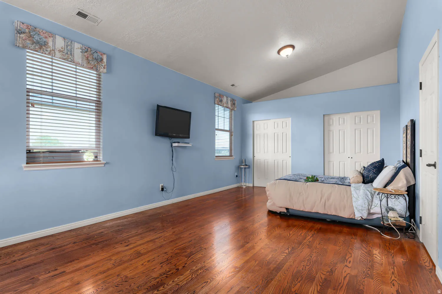 Spacious room featuring wood-finish flooring, two large windows with blinds, and a vaulted ceiling