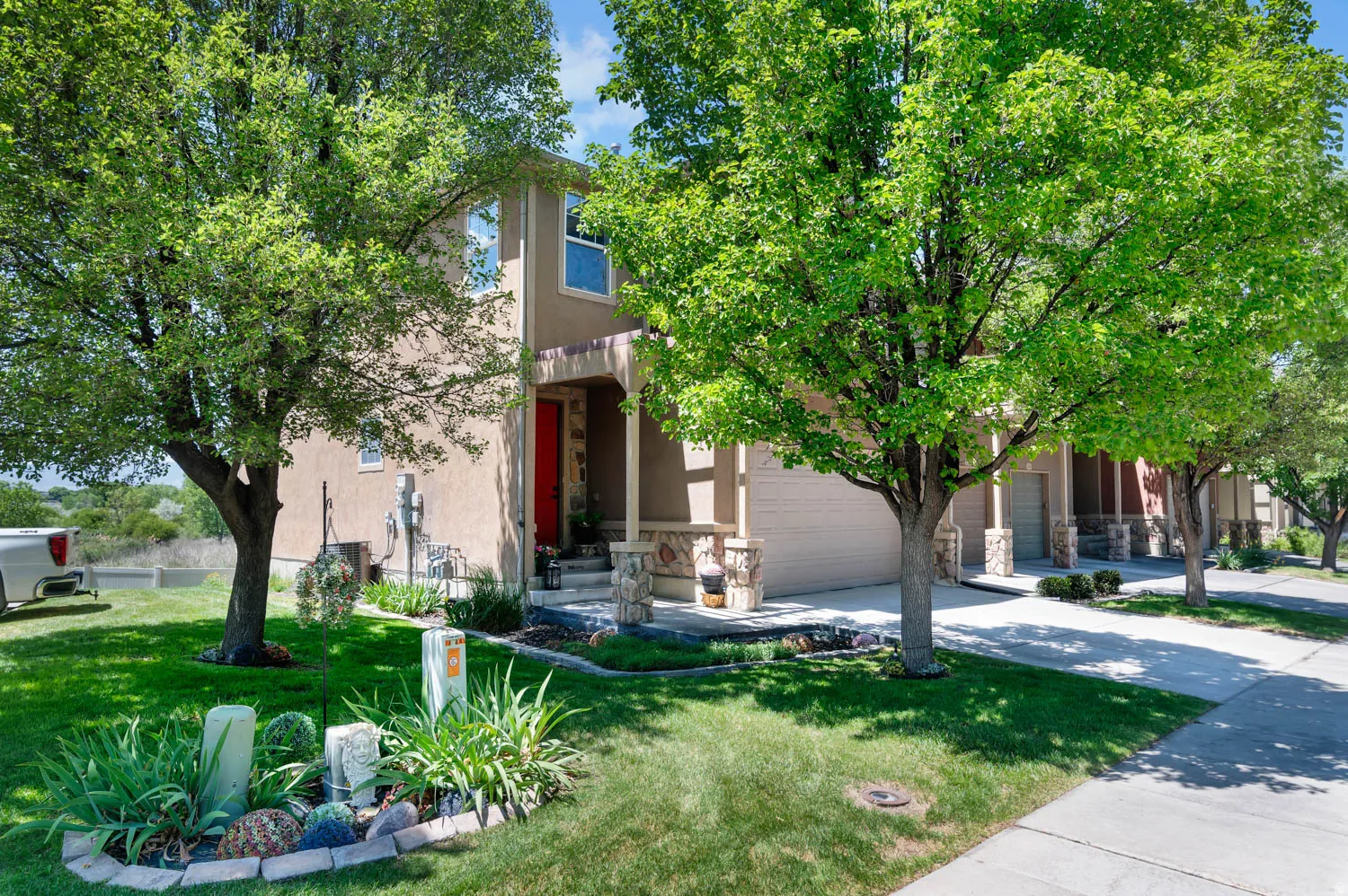 Exterior featuring a stucco finish, a covered porch with stone-clad columns, a red entry door, a two-car garage, and a manicured front lawn with mature trees
