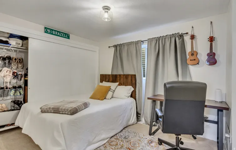 Bedroom featuring a sliding closet door, wood-finish headboard, window with privacy blinds and drapes, light-toned flooring, and a flush mount ceiling light