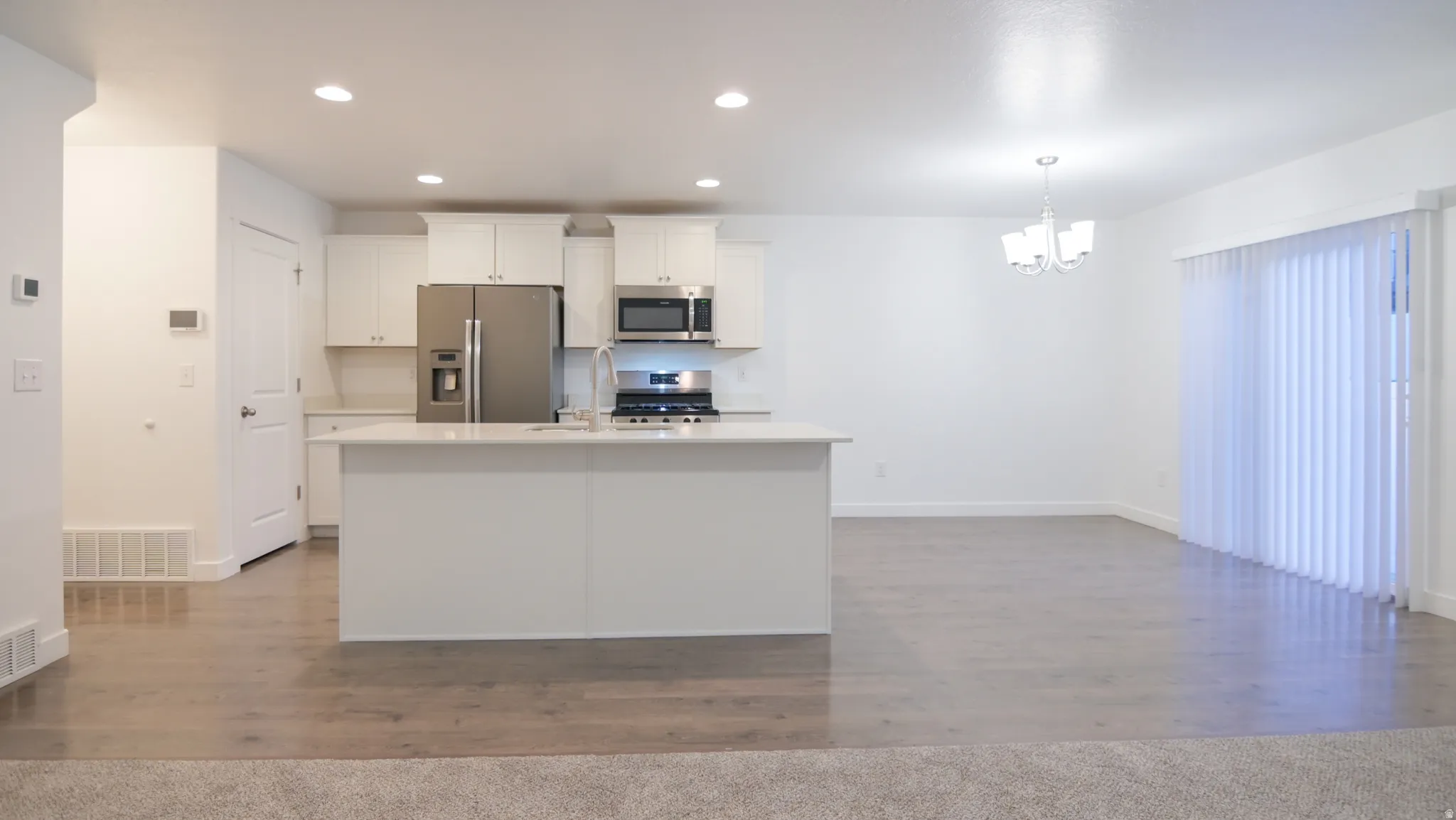 Open-concept living space featuring wood-finish flooring, a kitchen island with a sink, stainless steel appliances, white cabinetry, and a sliding glass door with vertical blinds