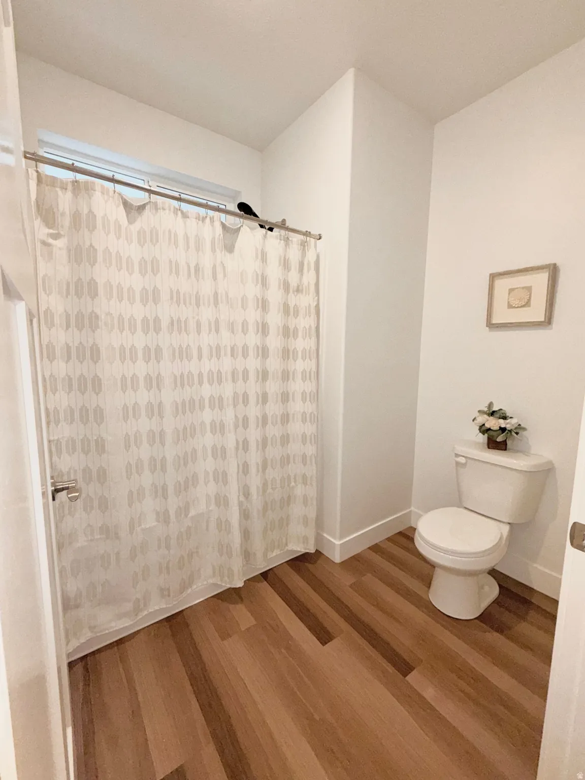 Bathroom featuring wood-finish flooring, a white toilet, and a shower area with a curtain rod