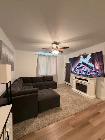 Living area featuring a ceiling fan, light-toned window treatments, and an electric fireplace unit