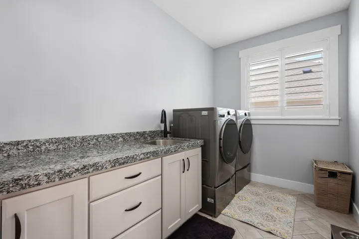 Laundry room featuring a built-in countertop with an undermount sink and matte black faucet, light-toned cabinetry with black hardware, and a window with white plantation shutters