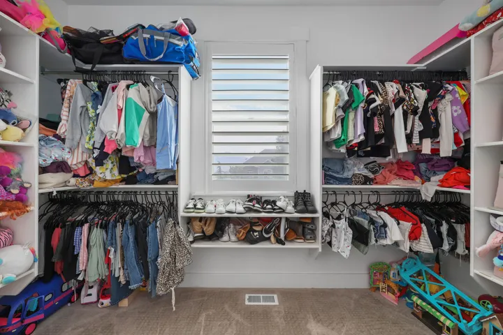 Walk-in closet featuring white built-in shelving, multiple hanging rods, a window with white louvered shutters, and light brown carpet flooring