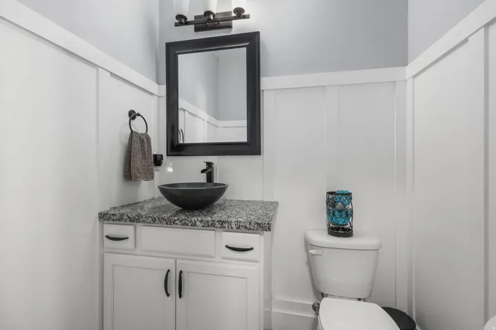 Powder room featuring board and batten wainscoting, a white vanity with black hardware, a vessel sink, a granite countertop, and an overhead light fixture