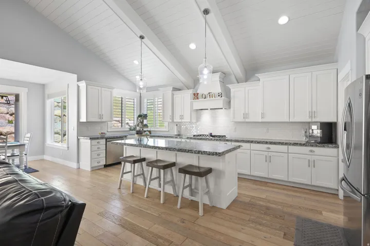 Open-concept kitchen featuring vaulted ceilings with exposed beams, white shaker cabinetry, granite countertops, and wood-finish flooring