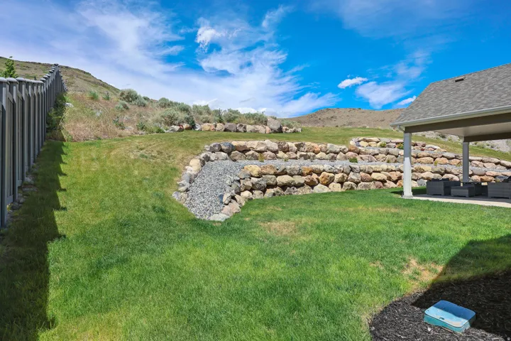 Expansive backyard featuring a covered patio with recessed lighting, tiered rock retaining walls, a gravel-lined swale, and a low-maintenance vinyl fence