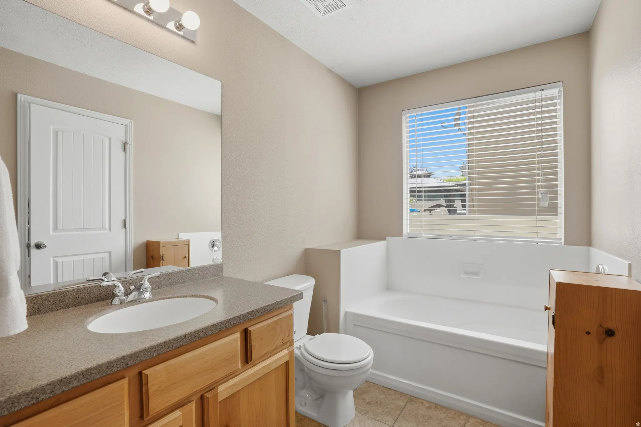 Bathroom featuring a built-in tub with surrounding ledge, a window with blinds, and a vanity with an undermount sink and stone-look countertop