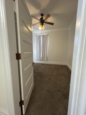 Spacious room featuring neutral carpeting, a ceiling fan with integrated lighting, window with light-filtering curtains, and classic crown molding
