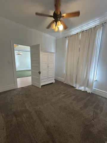 Carpeted room featuring a ceiling fan with integrated lighting, crown molding, wide baseboards, and a window with curtains