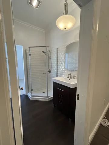 Bathroom featuring a dark wood-finish vanity with an integrated white sink, a corner glass-enclosed shower, white subway tile shower surround, and dark wood-finish flooring