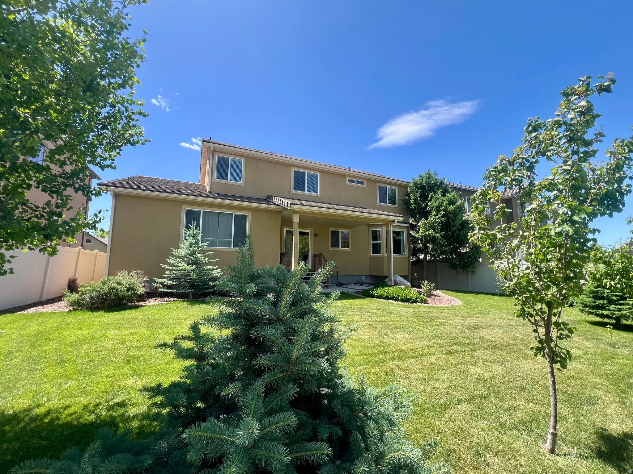 Expansive lawn featuring mature trees and shrubbery, a covered patio, and a two-story stucco exterior