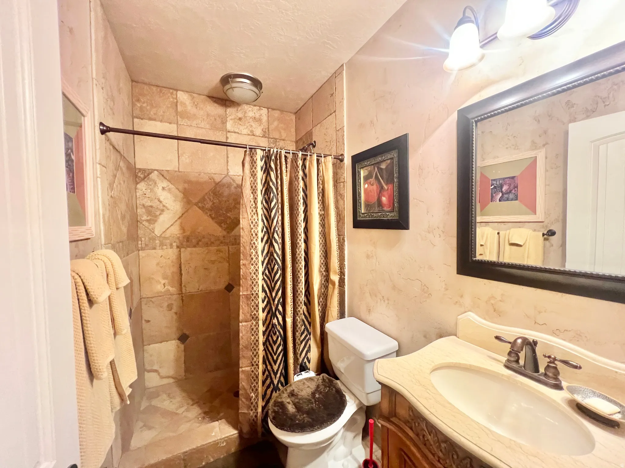 Bathroom featuring a walk-in shower with natural stone-look tile, a single vanity with an integrated sink and bronze-finish faucet, and a decorative framed mirror