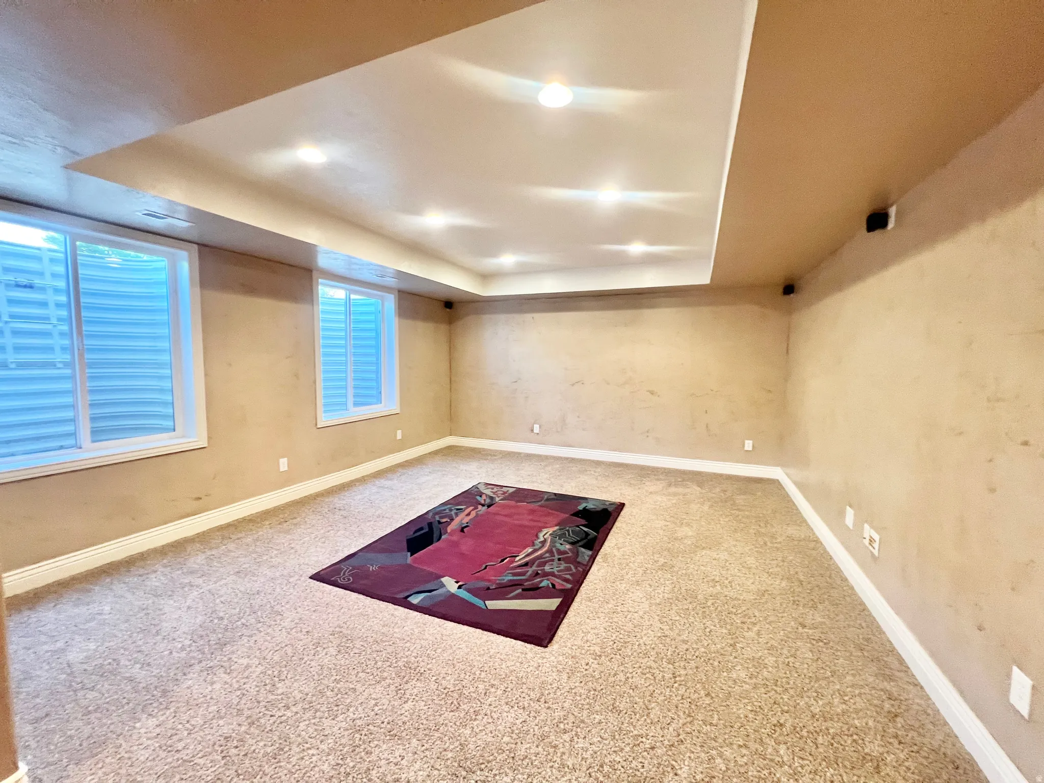 Recessed ceiling with integrated lighting, two egress windows, wall-mounted speakers, and neutral carpet flooring
