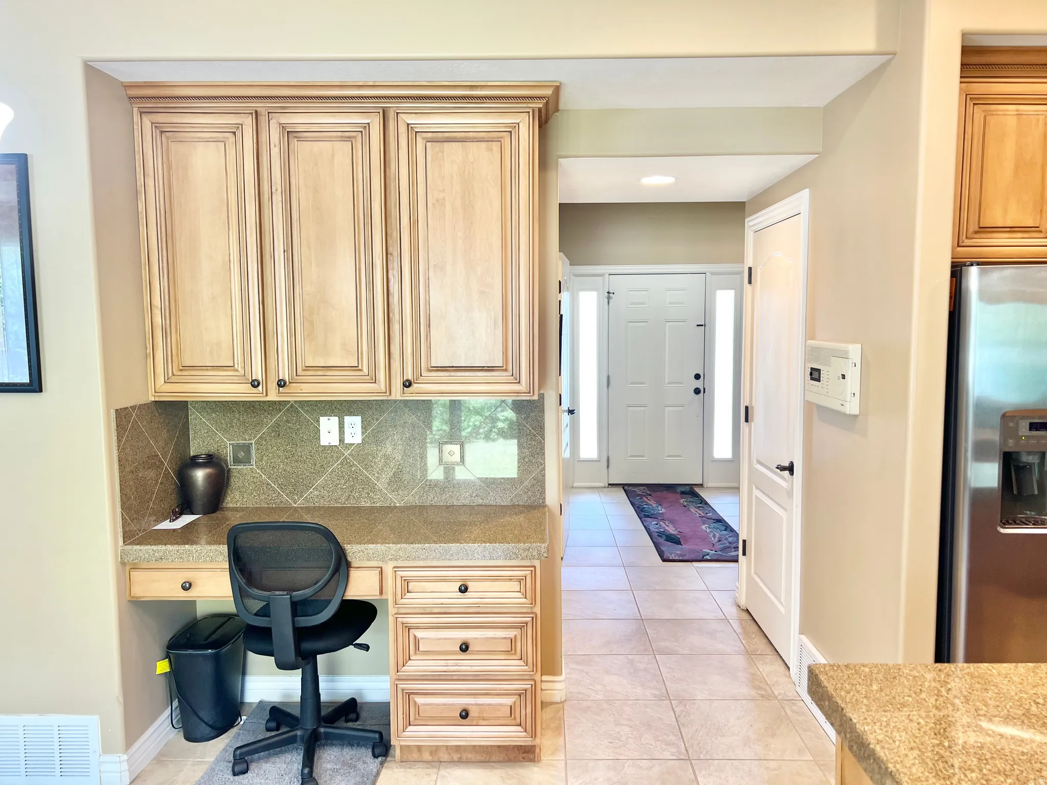 Built-in desk area featuring light wood cabinetry with upper storage, a stone-look countertop, and a tiled backsplash
