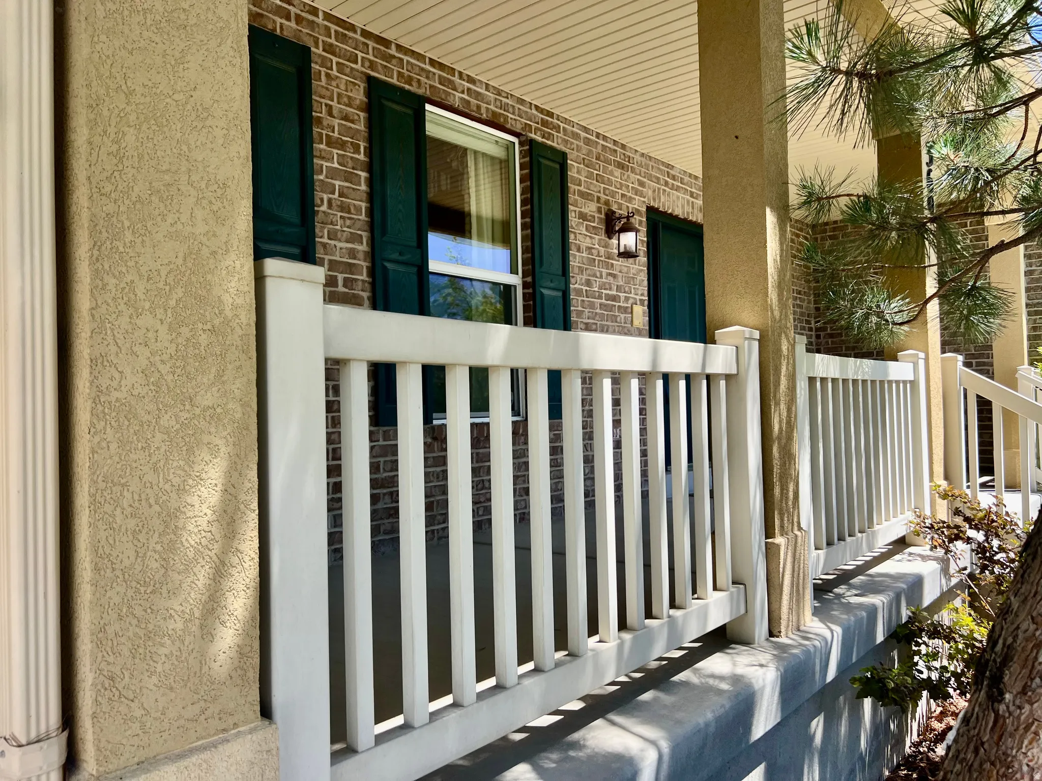 Expansive front porch featuring white railing, textured stucco columns, brick facade, and dark green louvered shutters