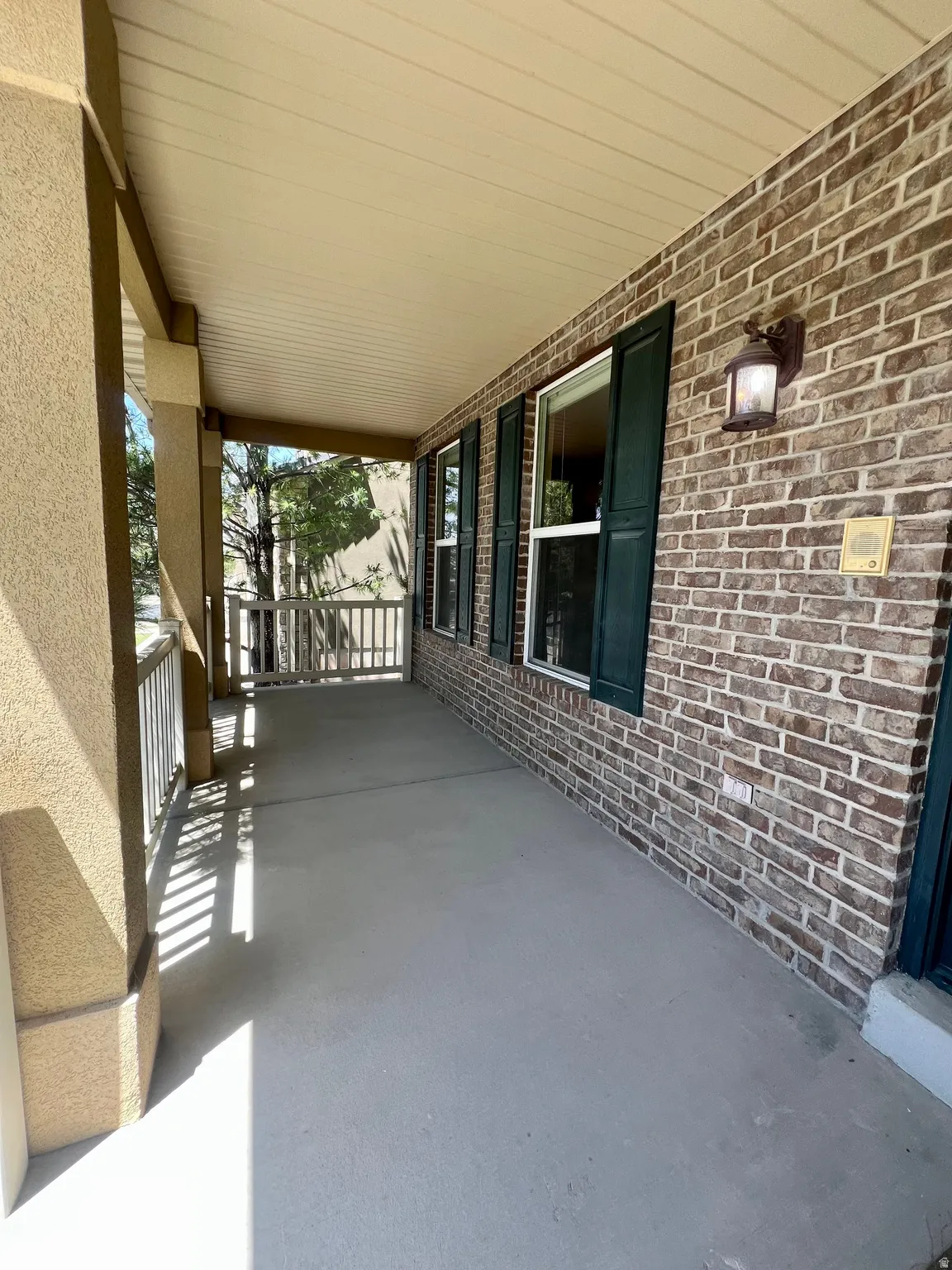 Covered front porch featuring a brick facade, decorative wall sconce, and dark green louvered shutters
