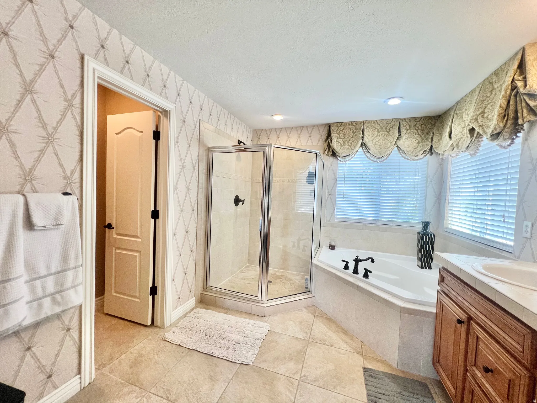 Spacious bathroom featuring a corner soaking tub, glass-enclosed shower, wood vanity with integrated sink, tiled flooring, and multiple windows with blinds