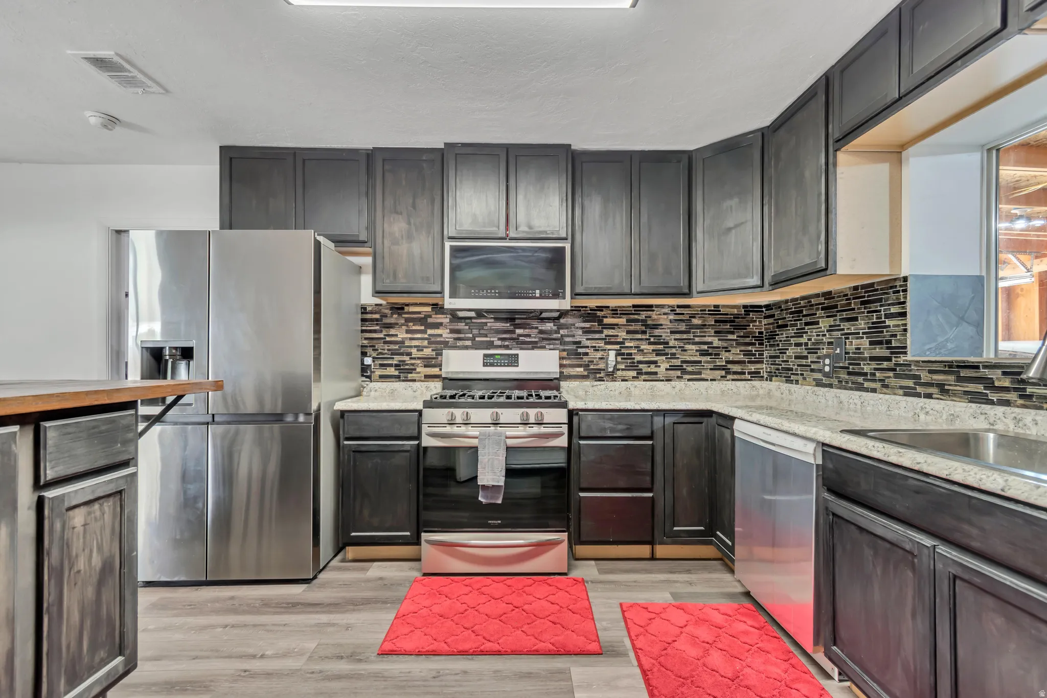 Kitchen featuring dark wood-finish cabinetry, stainless steel appliances, a multi-tone tile backsplash, light-toned countertops, and wood-finish flooring