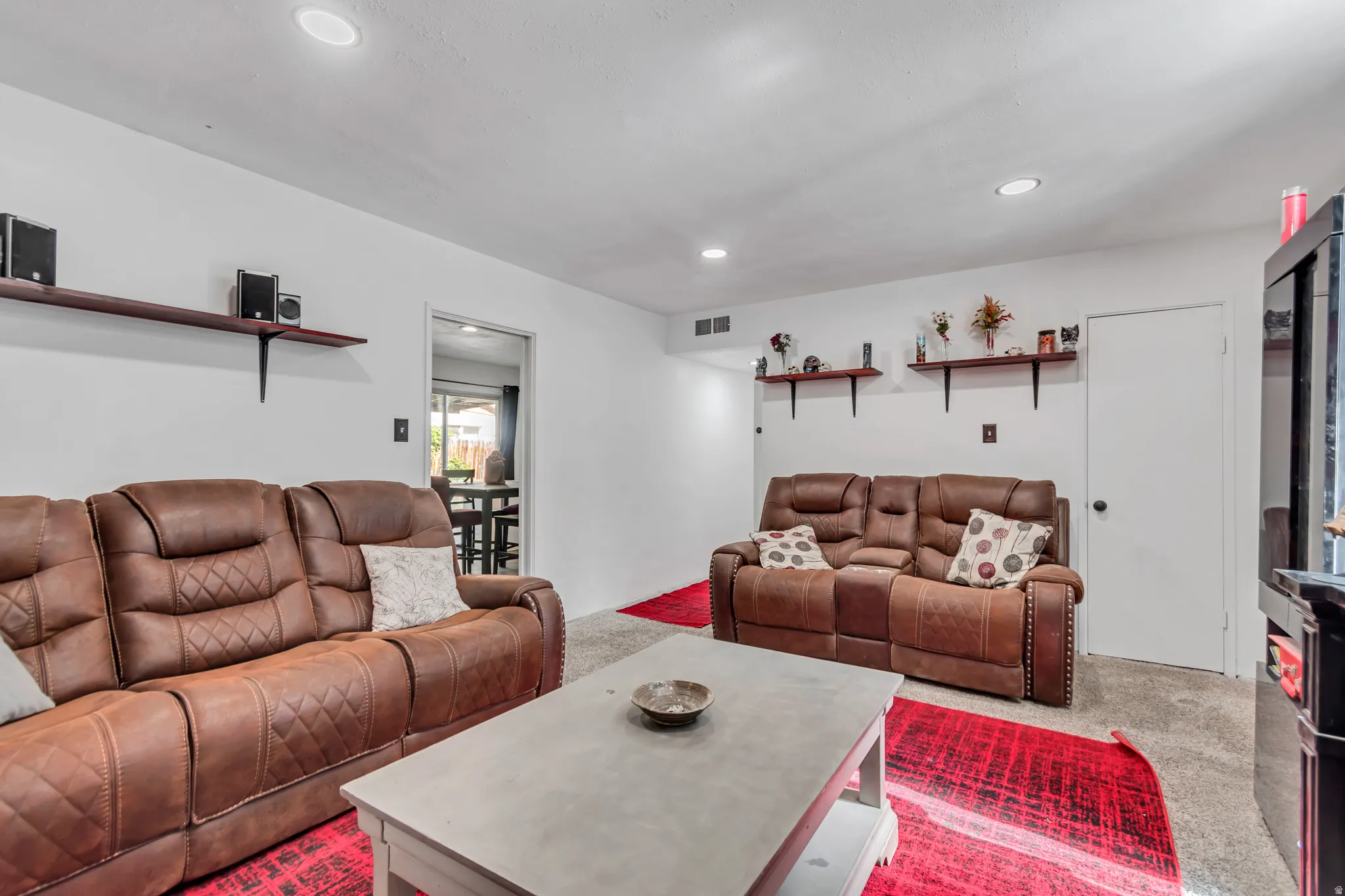 Recessed lighting illuminates this living space, featuring wall-mounted shelving and light-colored carpeting