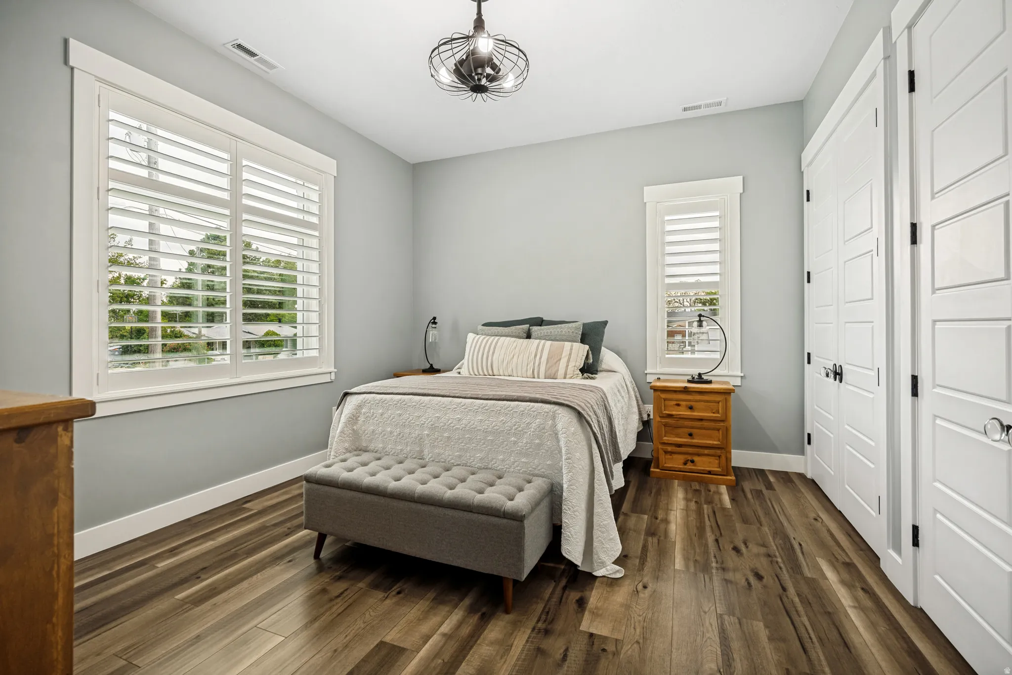 Bedroom featuring wood-finish flooring, two windows with white plantation shutters, and a modern ceiling fixture