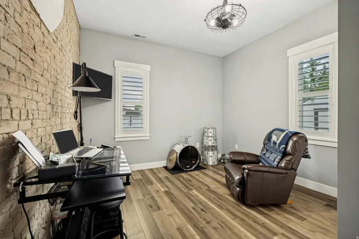 Room featuring a prominent exposed brick wall, wood-finish flooring, two windows with plantation shutters, light gray painted walls, and a contemporary ceiling fixture