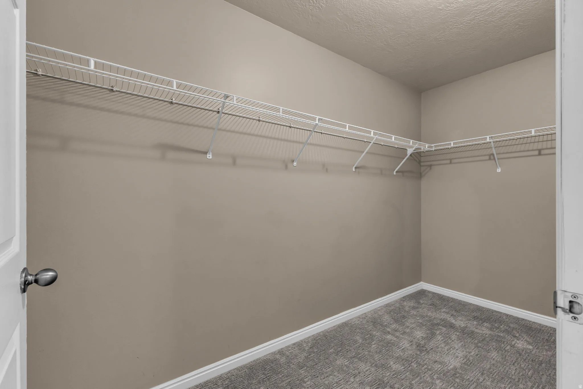 Spacious walk-in closet featuring light beige walls, white wire shelving, and grey carpet flooring