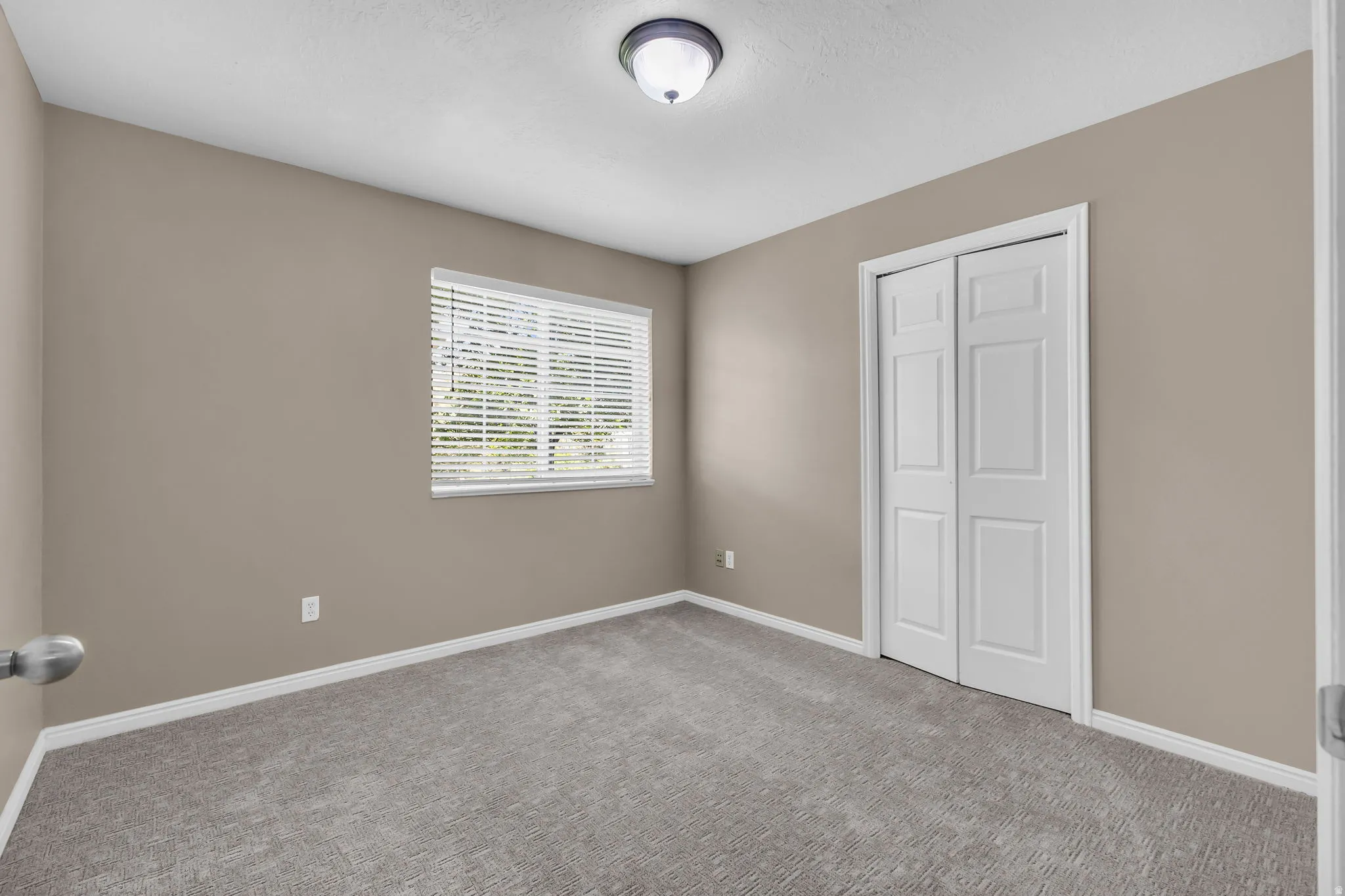 Neutral-toned room featuring a single window with blinds, carpet flooring, a flush-mount ceiling light fixture, and a bi-fold closet door