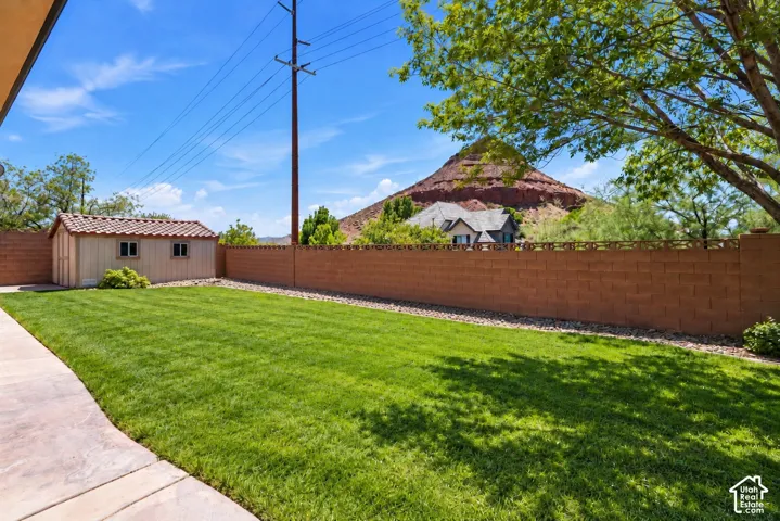 Backyard featuring a manicured lawn, concrete patio, masonry perimeter wall, detached storage structure with tile roofing, and prominent mountain backdrop