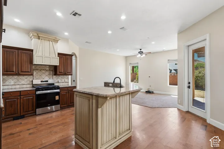 Kitchen featuring wood-finish flooring, dark wood cabinetry, a decorative range hood, stainless steel stove, and a central island with a sink and light-toned countertop