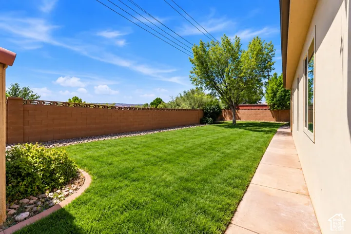 Backyard featuring a manicured lawn, concrete perimeter walkway, masonry block wall fencing, river rock landscaping, and mature trees