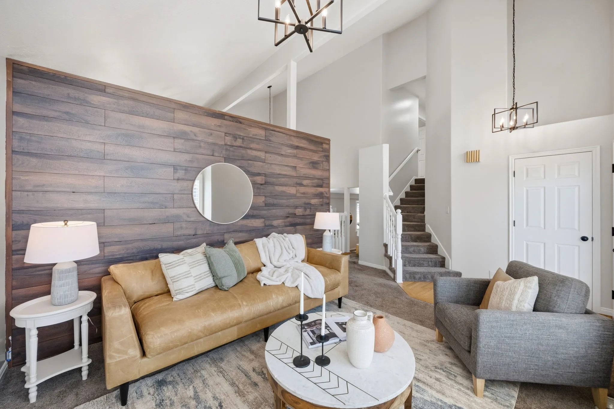 Vaulted ceiling living room featuring a wood-panel accent wall, contemporary lighting fixtures, light-toned carpeting, and a staircase with white railings
