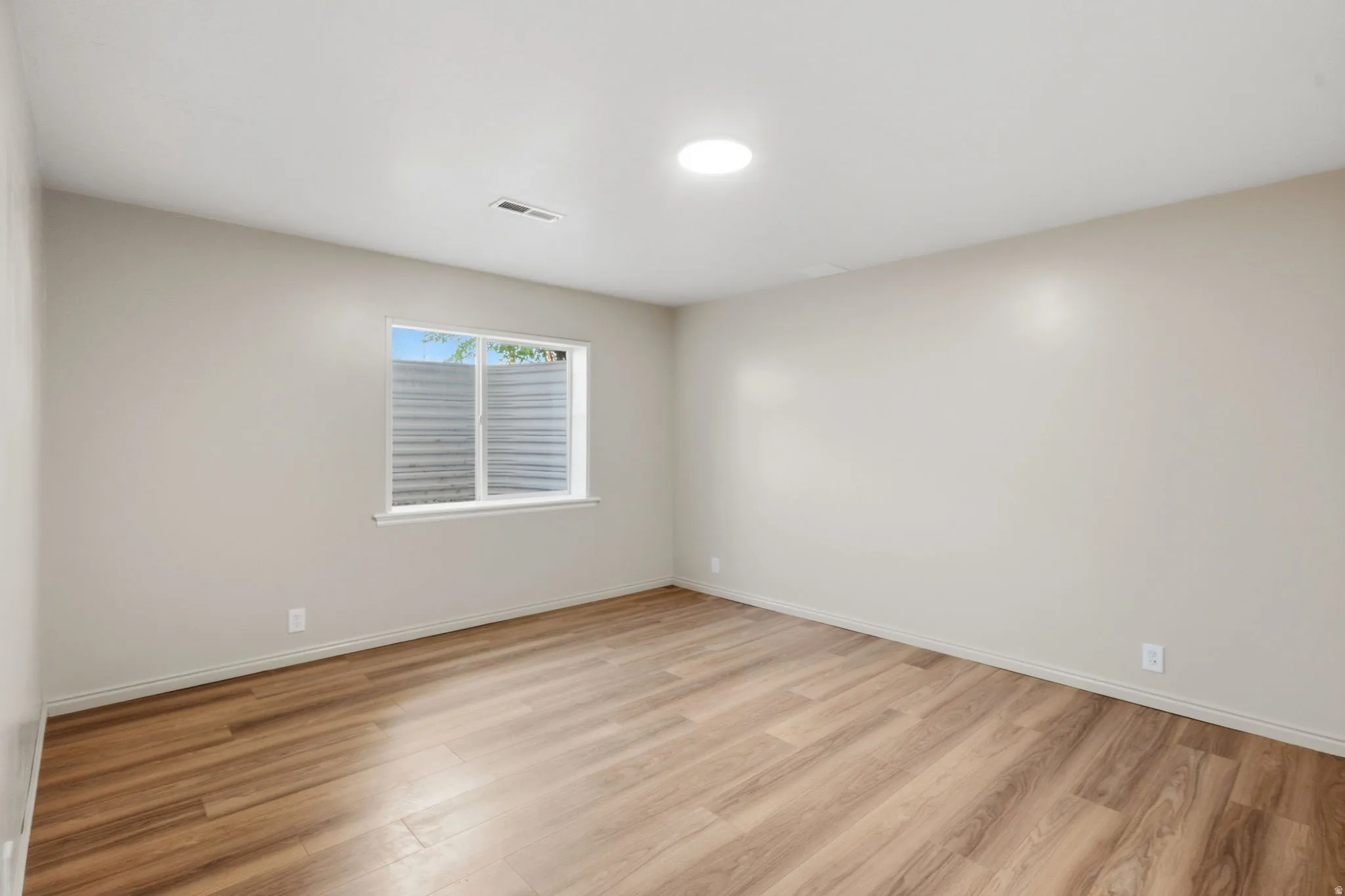 Spacious room featuring wood-finish flooring, a window with exterior grating, a recessed ceiling light, and light-toned wall finishes