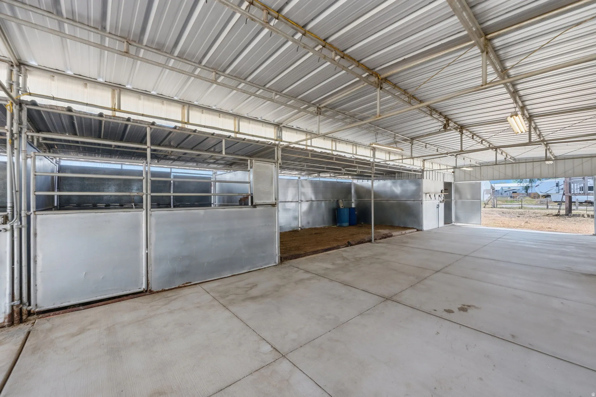 Covered equestrian facility featuring multiple stalls with metal gates, an open concrete aisle, and a corrugated metal roof structure